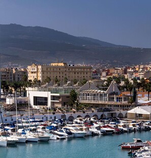 A marina with boats, a modern building with a glass facade, and a hilly cityscape in the background. - Melilla Puerto Hotel (Melilla)