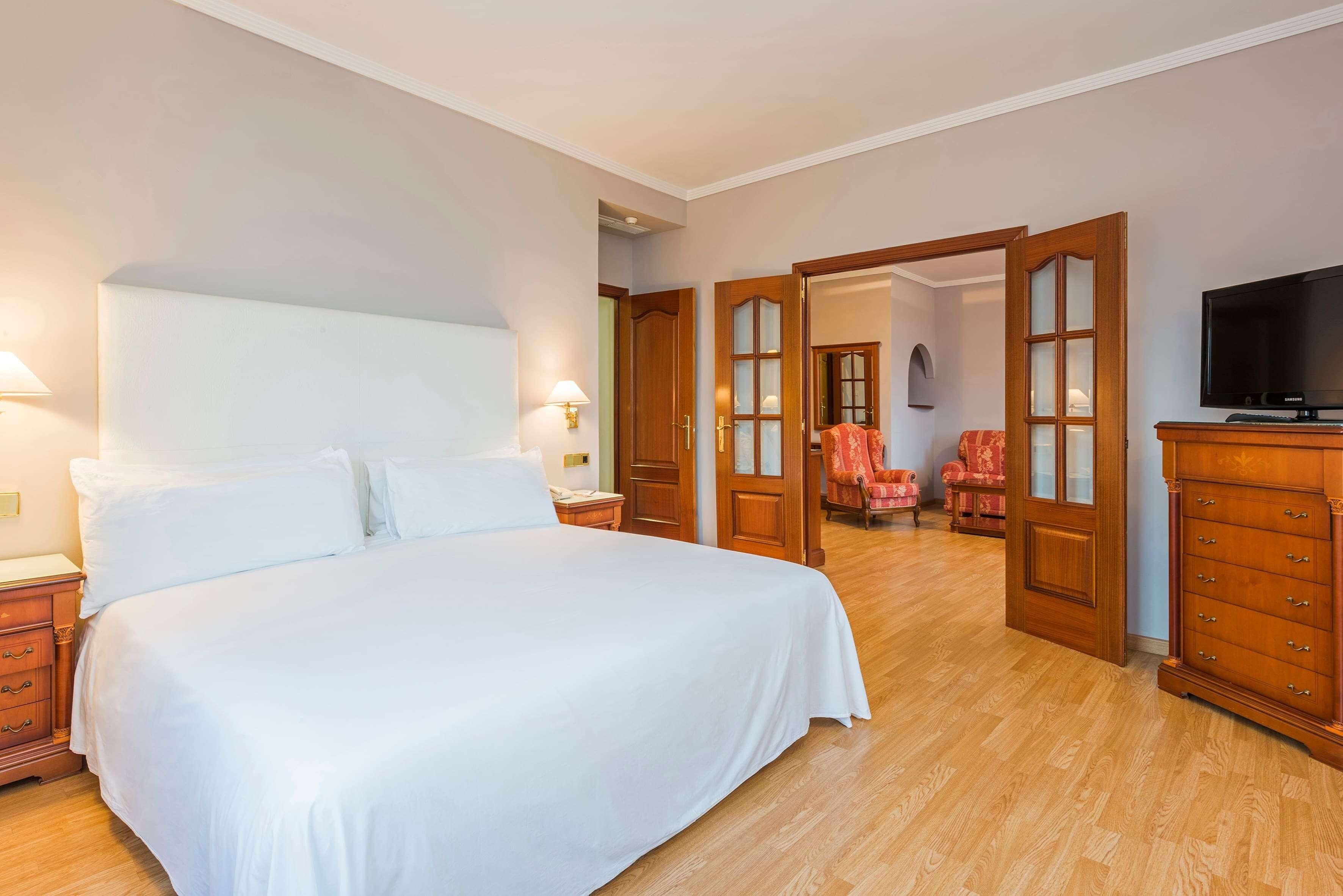 Un dormitorio con una cama grande, cómoda de madera, televisión y una silla al fondo.