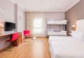 A hotel room with a bunk bed, a desk with a TV, a red chair, a small table, and a bedside lamp.