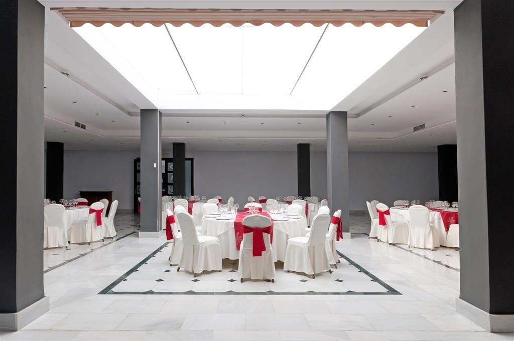 A spacious hall with round tables and white chairs, a central stage, and a high ceiling with skylights.