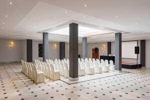 Un amplio salón con filas de sillas blancas, un escenario y una pantalla para proyector.