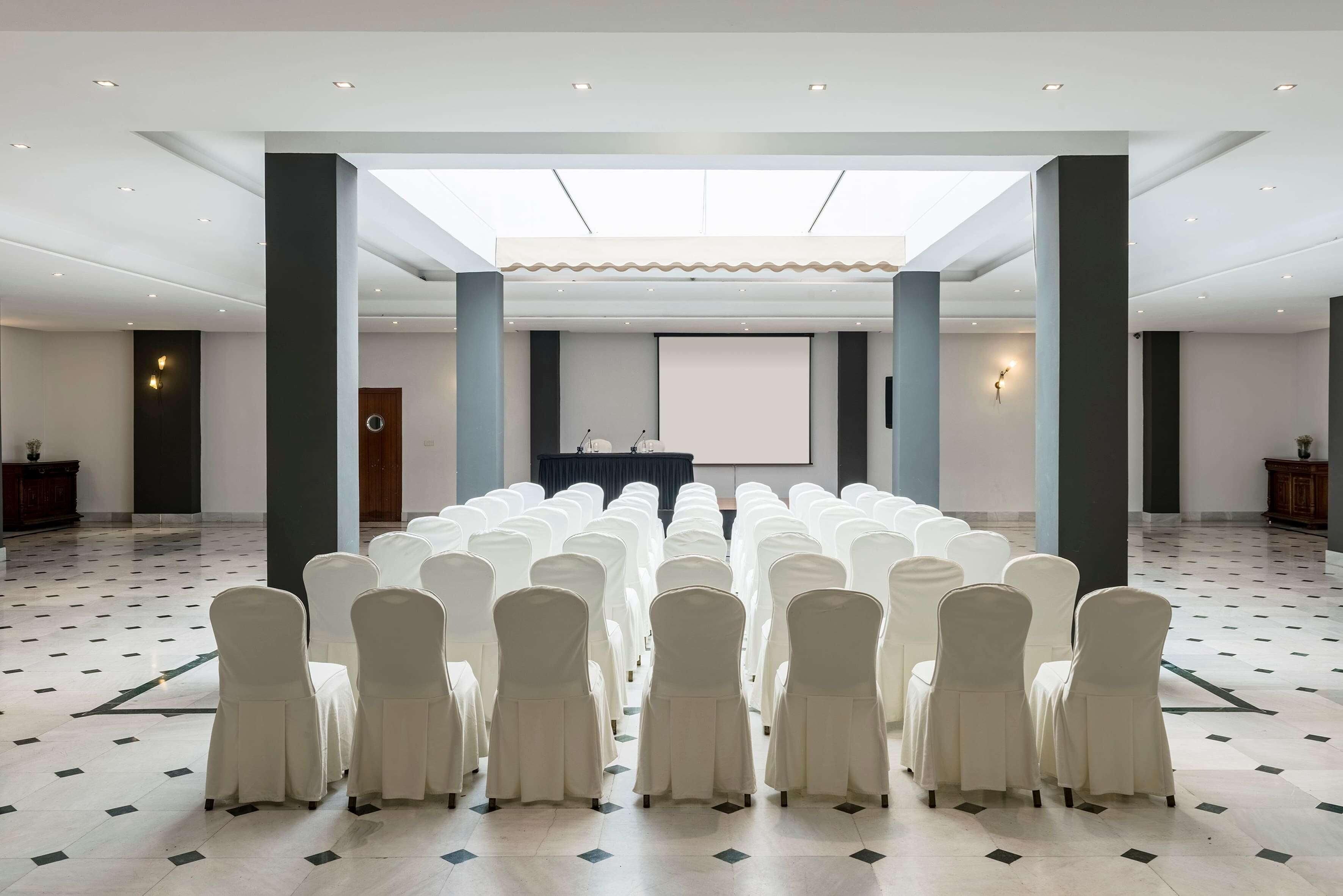 Une salle de conférence avec des rangées de chaises blanches, un pupitre et un écran de projection.