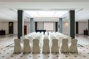 Una sala de conferències amb files de cadires blanques, un escenari i una pantalla projectora.
