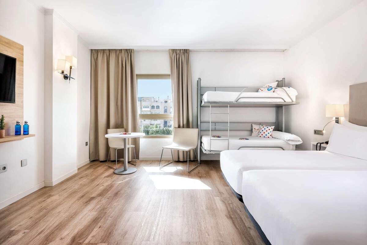 Una camera d'albergo con un letto a castello, una scrivania, una sedia, un tavolino e una televisione.