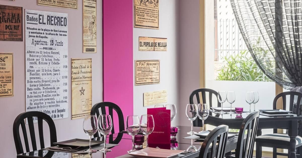 Interior de un restaurante con una pared de acento rosa, varios menús a la vista y una zona de comedor con mesas dispuestas para los clientes.