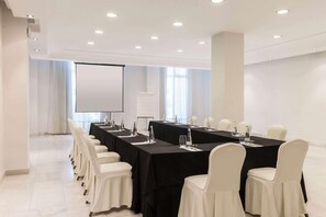 Un salón de conferencias con una larga mesa, sillas blancas y una pantalla de proyector.