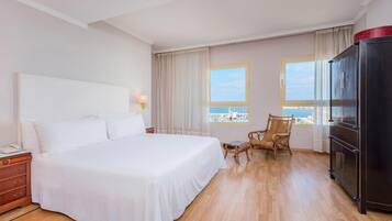 Habitación de hotel con una cama grande, televisión, armario, escritorio con lámpara y vista al mar.