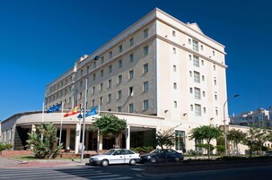 Un hotel de varios pisos con una entrada curva, exhibición de banderas y un estacionamiento.