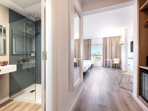 Quarto de hotel com chuveiro em gabinete de vidro, cama, secretária, cadeira, televisão e vista para a cidade.