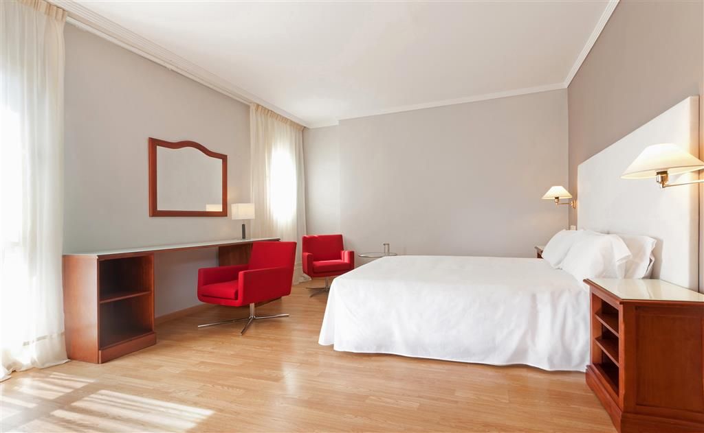 A hotel room with a bed, a desk, two red chairs, a wooden bookshelf, and a large mirror.