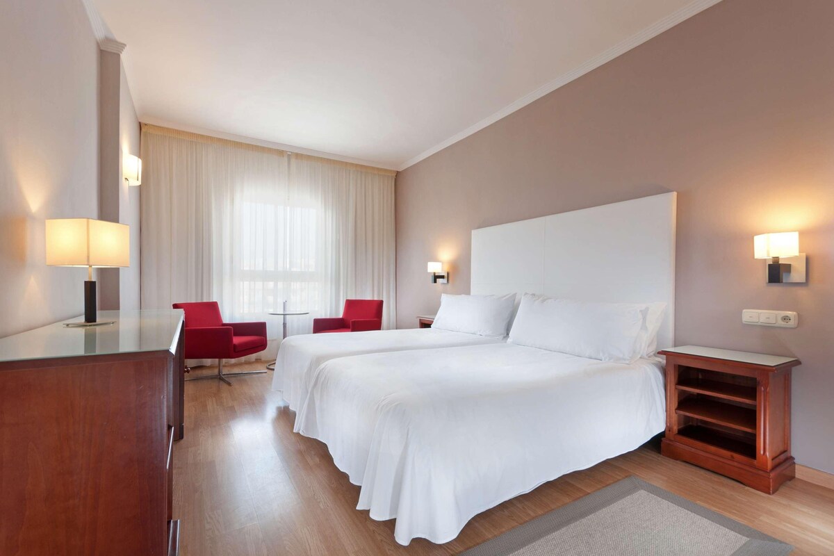 Camera d'albergo con due letti, una scrivania, una sedia rossa, un tavolino e un'ampia finestra con tende.