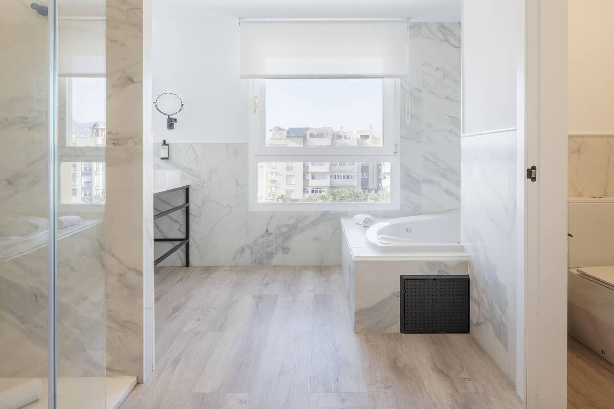 Une salle de bain moderne équipée d’une baignoire, d’une douche et d’une fenêtre donnant sur des immeubles.