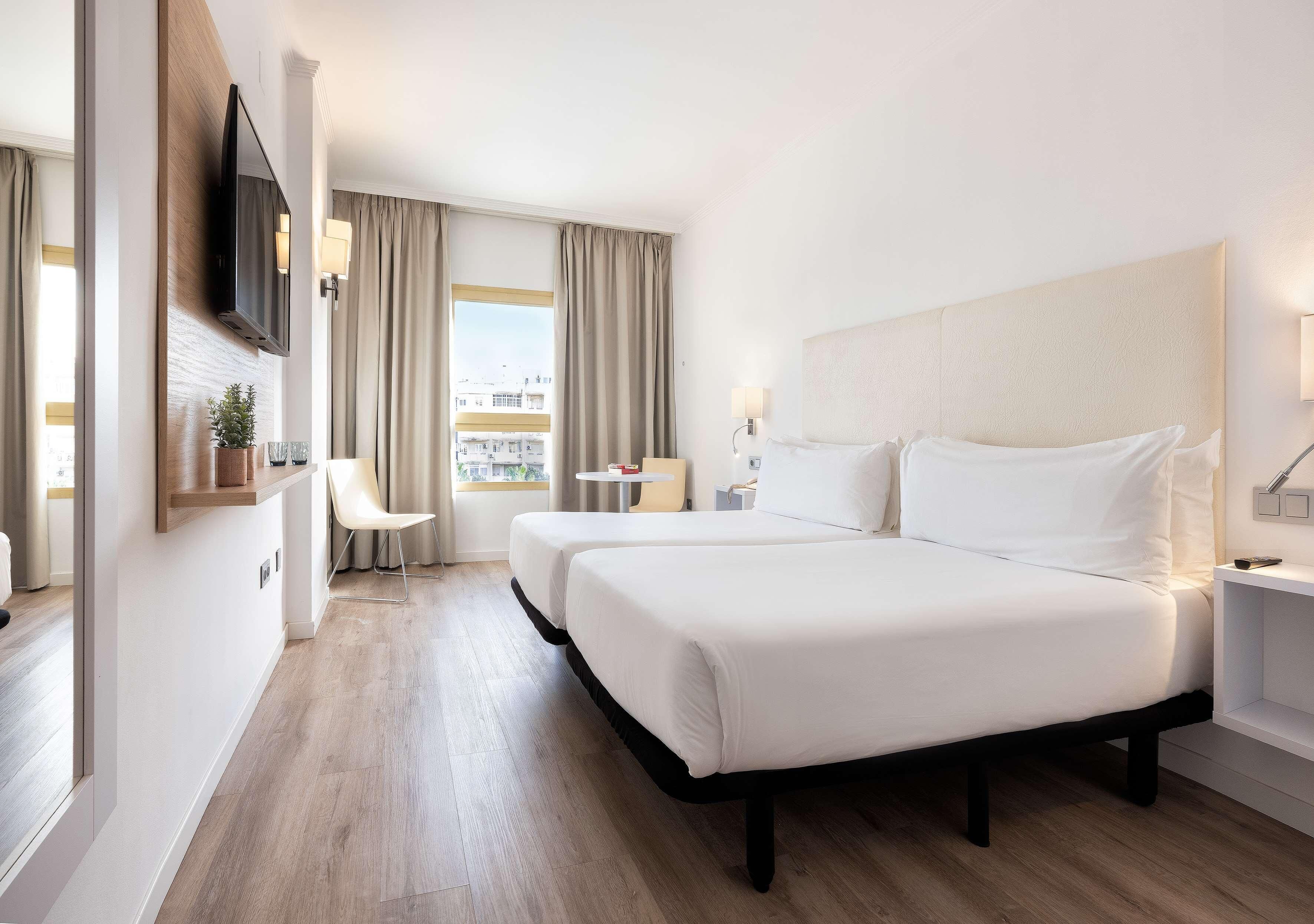 Une chambre d’hôtel avec deux lits, une télévision, une petite table, une chaise et une fenêtre avec des rideaux.