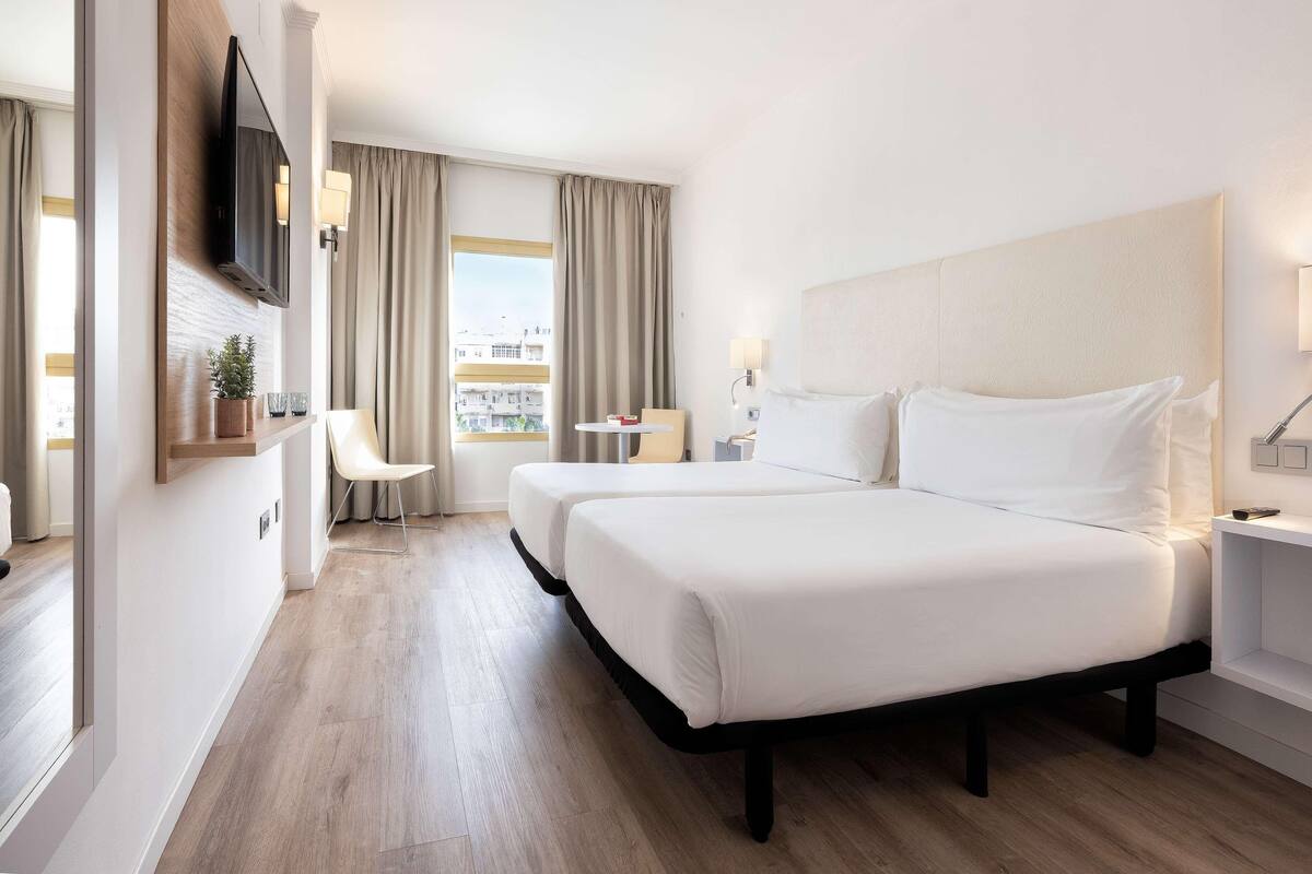 A hotel room with two beds, a TV, a small table, a chair, and a window with curtains.