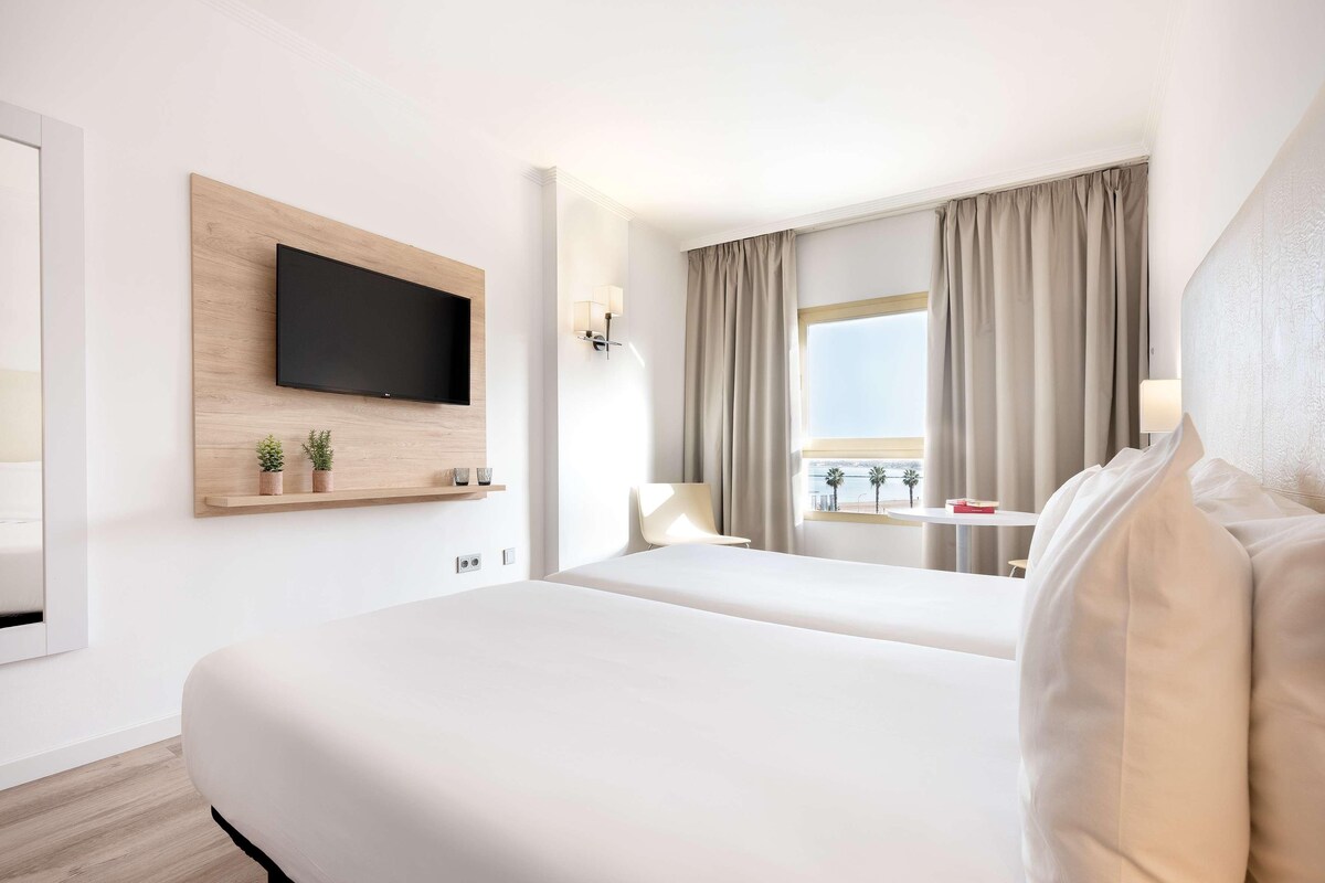 Camera d'albergo con un letto, una TV montata su un pannello di legno, una finestra con tende e vista su palme.