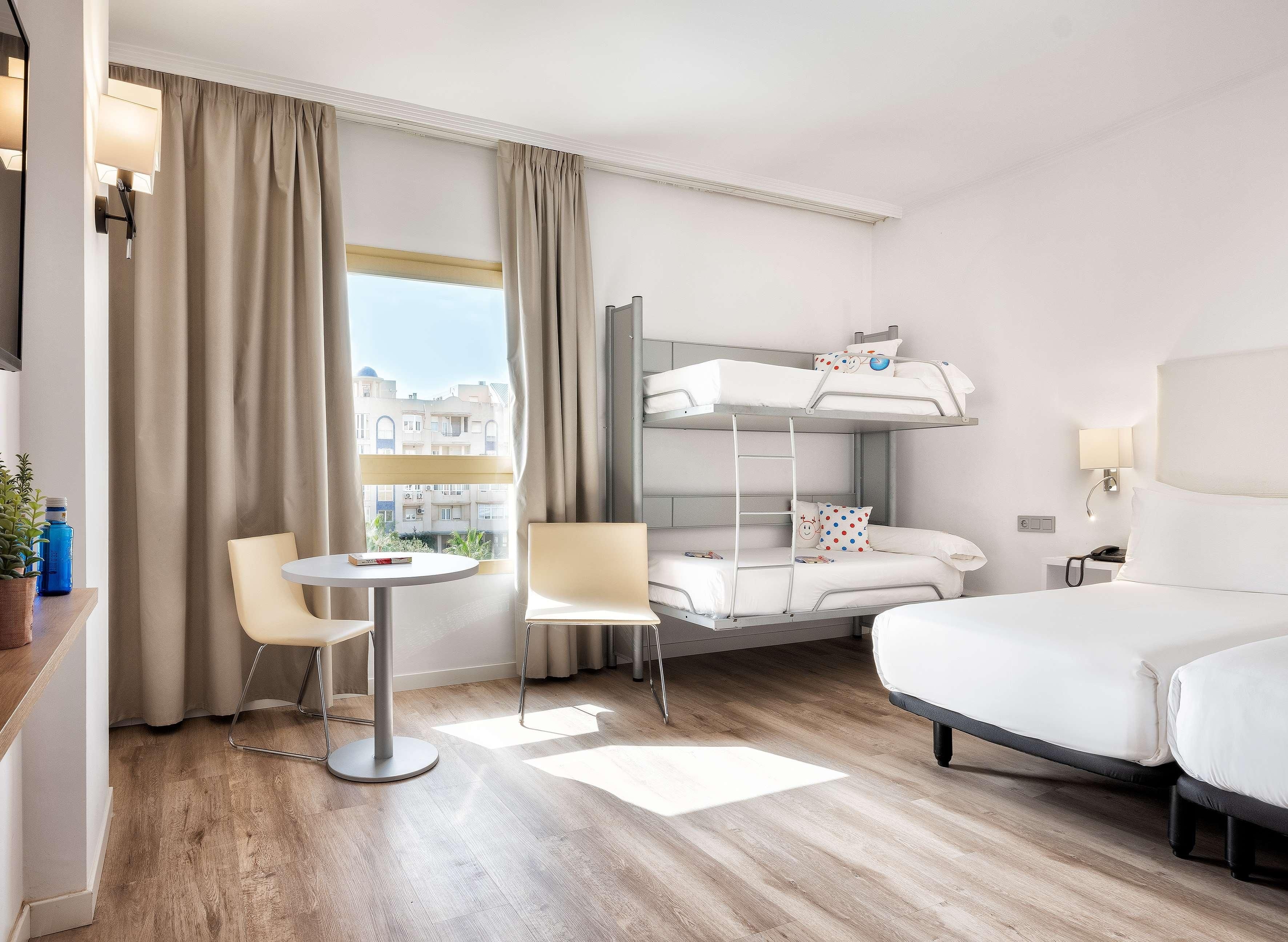 Habitación de hotel con literas, una cama individual, una mesa redonda, dos sillas, una mesita de noche, una lámpara y vistas a edificios.