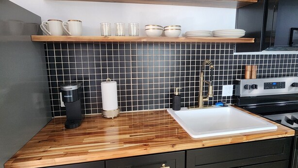 Una cocina moderna con armarios negros, encimera de madera y salpicadero de azulejos negros. Hay dos estantes sobre la encimera con varios utensilios de cocina.