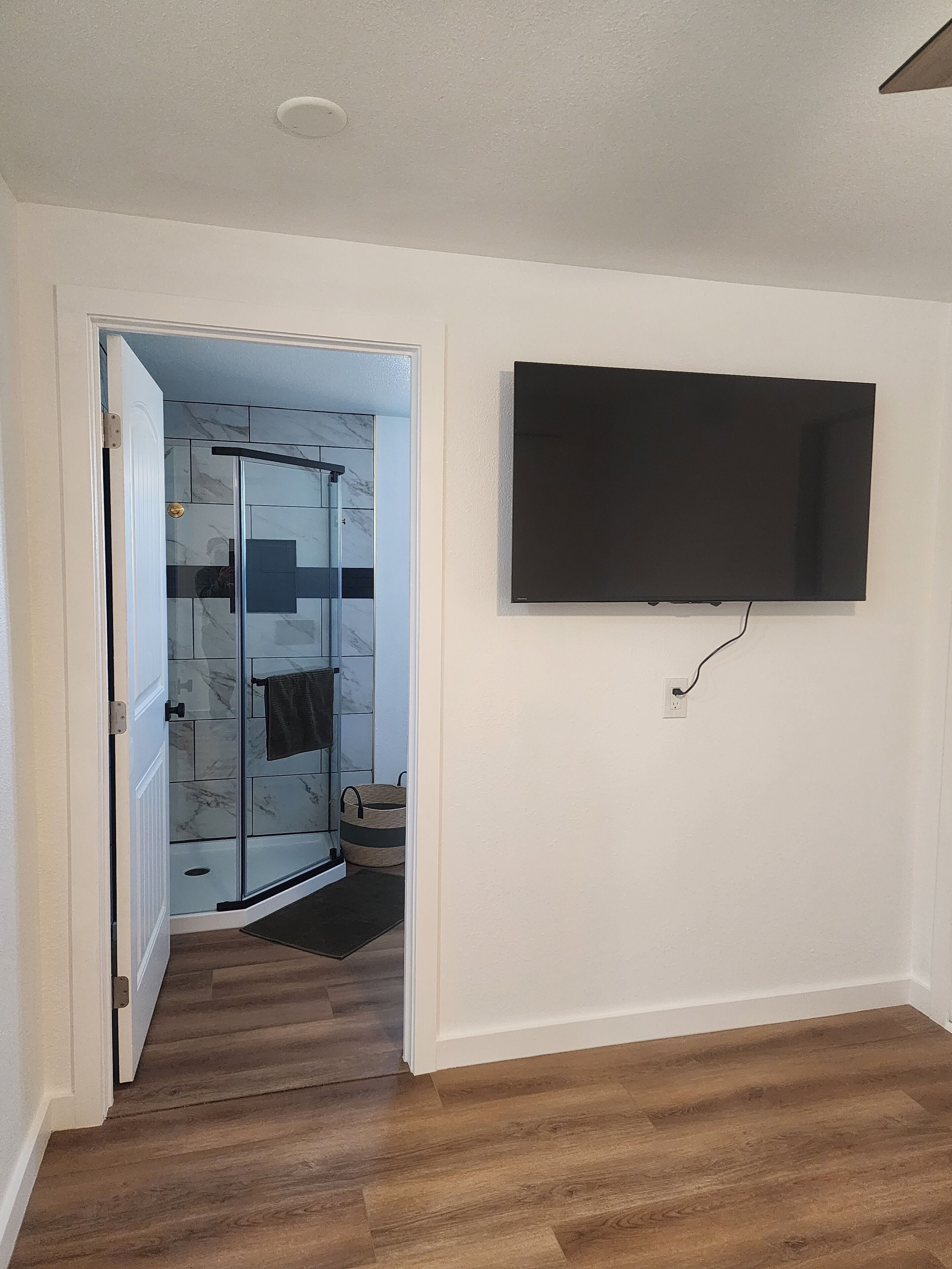 Quarto com piso de madeira, box de vidro, televisão montada e porta que dá para outro ambiente.