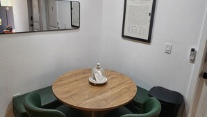 Un pequeño comedor con una mesa redonda de madera, cuatro sillas tapizadas en verde, un reloj de pared, un menú enmarcado y un cesto de basura.
