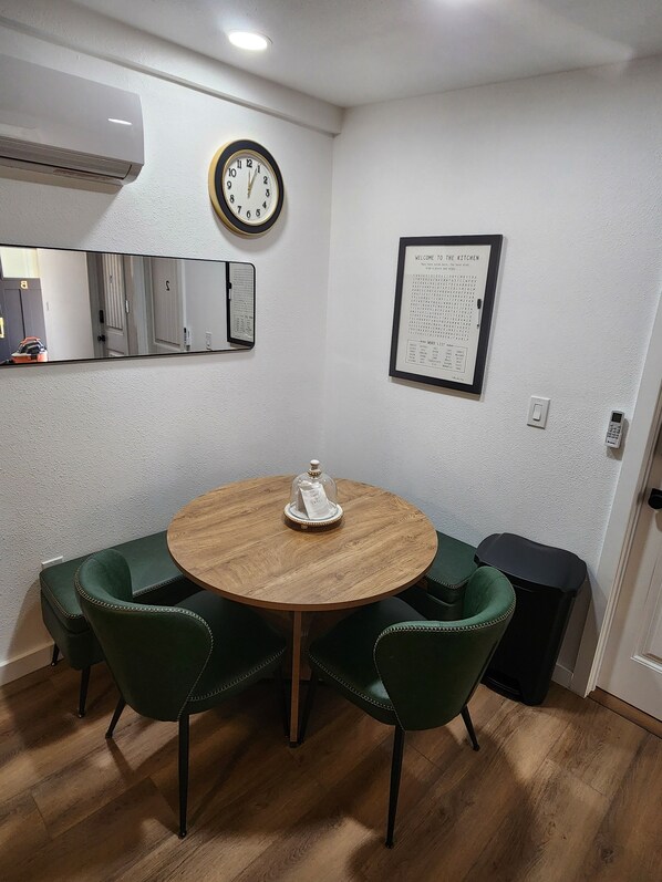 A small dining area with a round wooden table, four green upholstered chairs, a wall clock, a framed menu, and a trash bin.