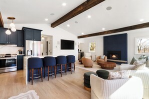 Une cuisine moderne avec des meubles de couleur sombre, un îlot central doté de tabourets de bar, un coin repas avec une table basse ronde et un salon avec une cheminée.