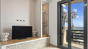 Una sala de estar moderna con un televisor de pantalla plana, un banco con textura de piedra, objetos decorativos y un puerta corrediza de vidrio que da a un balcón con vistas a palmeras y un edificio con el rótulo "SUMAR".