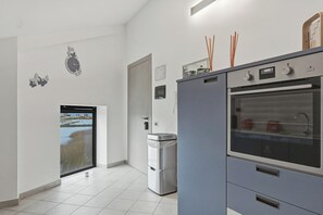 A modern kitchen with a built-in oven, a refrigerator, and a window with a view of a body of water. - Case Vacanze Celeste (Marzamemi)