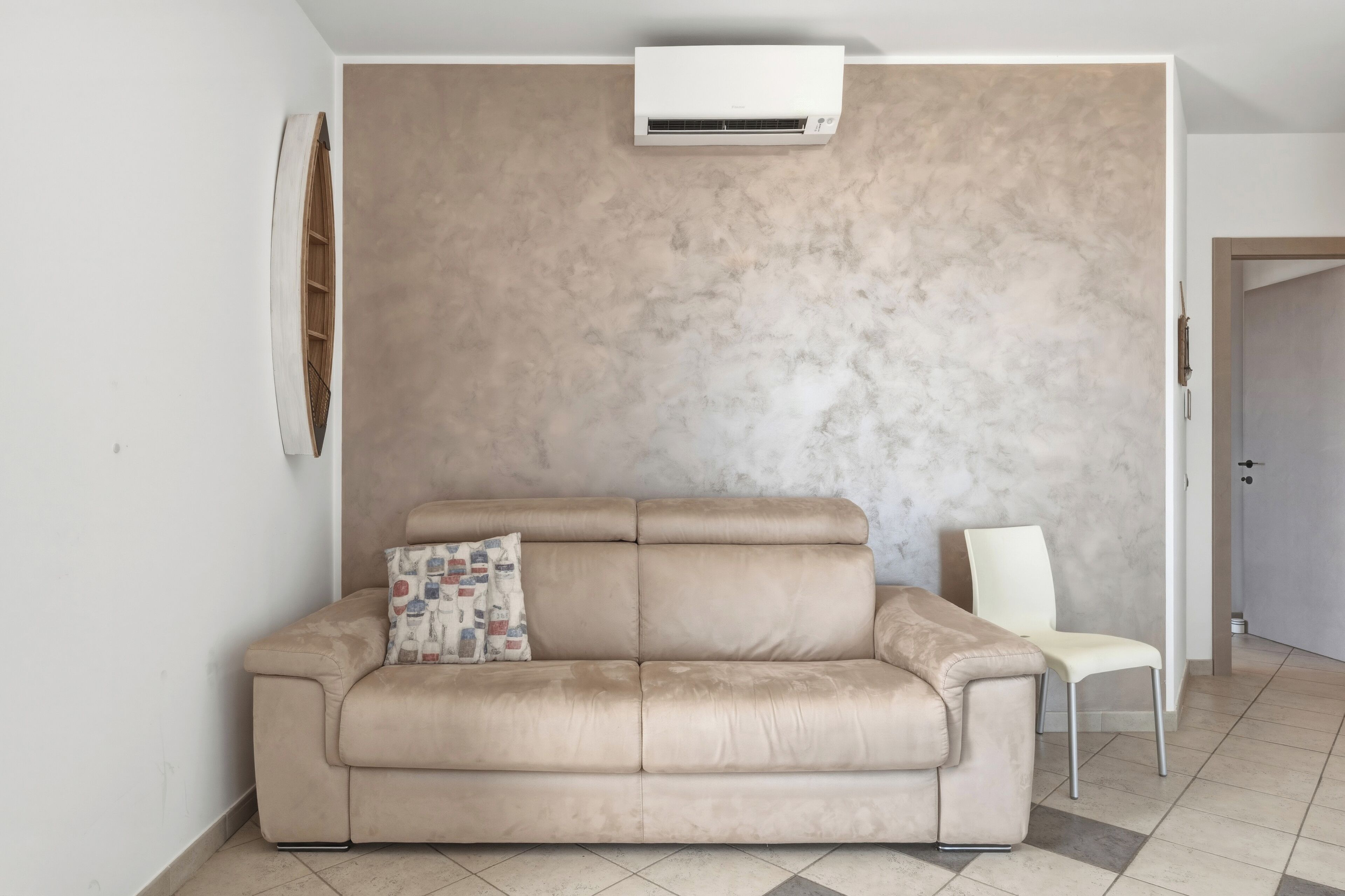 Ein beiges Ledersofa mit gemustertem Kissen, ein weißer Sessel und eine wandmontierte Klimaanlage in einem Raum mit strukturierter beiger Wand und Fliesenboden.