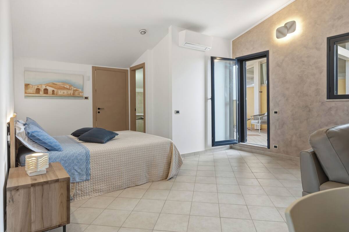 A bedroom with a bed, a nightstand, a chair, a painting on the wall, and a door leading to a balcony.