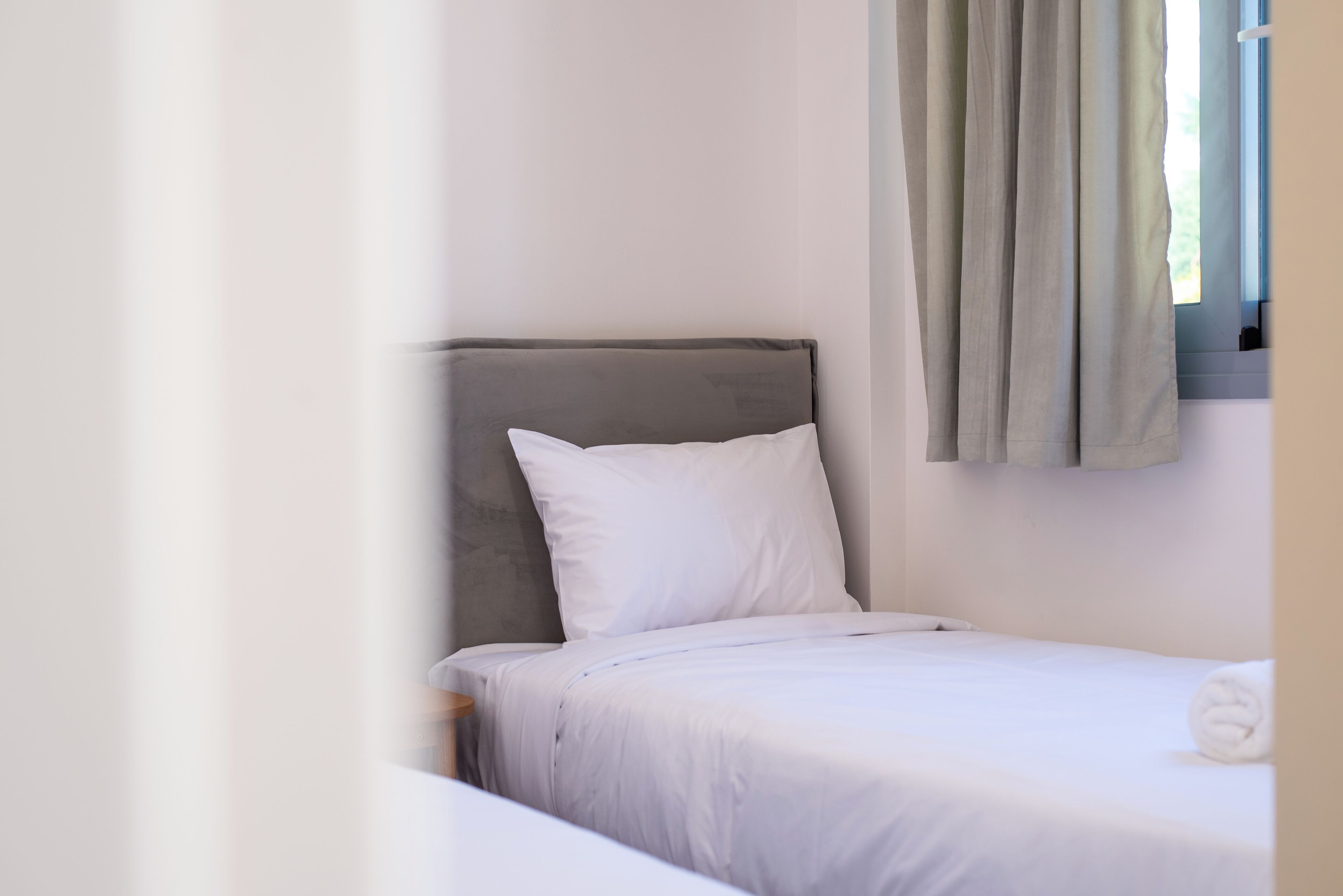 A single bed with a grey headboard, white bedding, and a window with curtains.