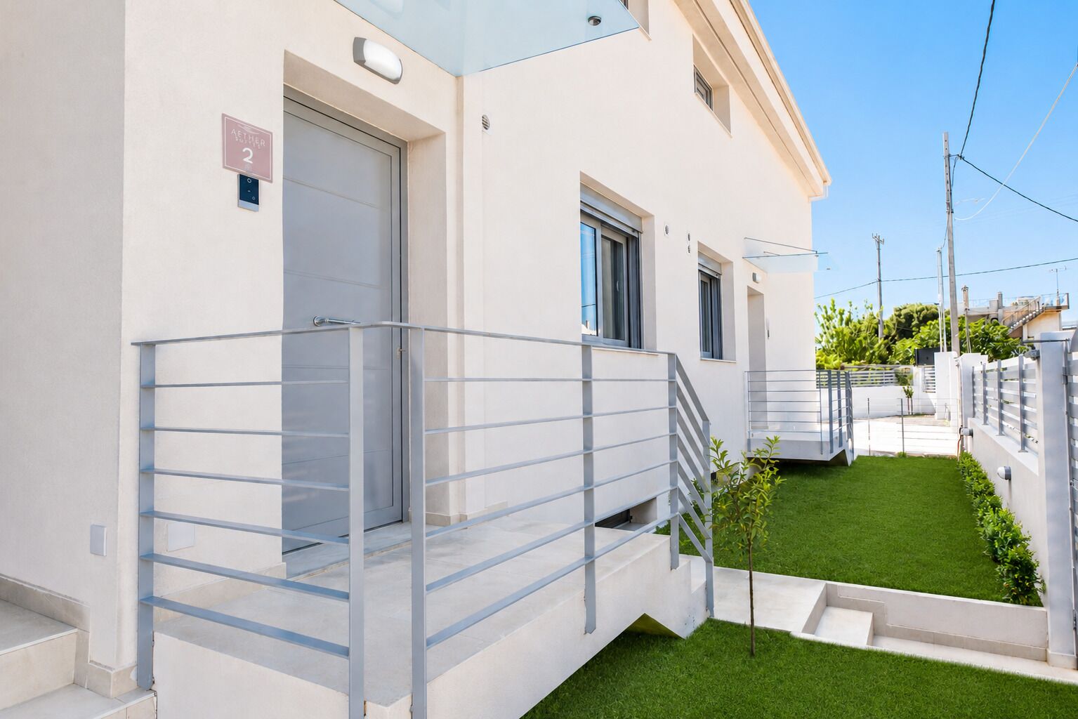 Un edificio residencial moderno con balcón de vidrio transparente, un pequeño césped y una puerta visible con un número.