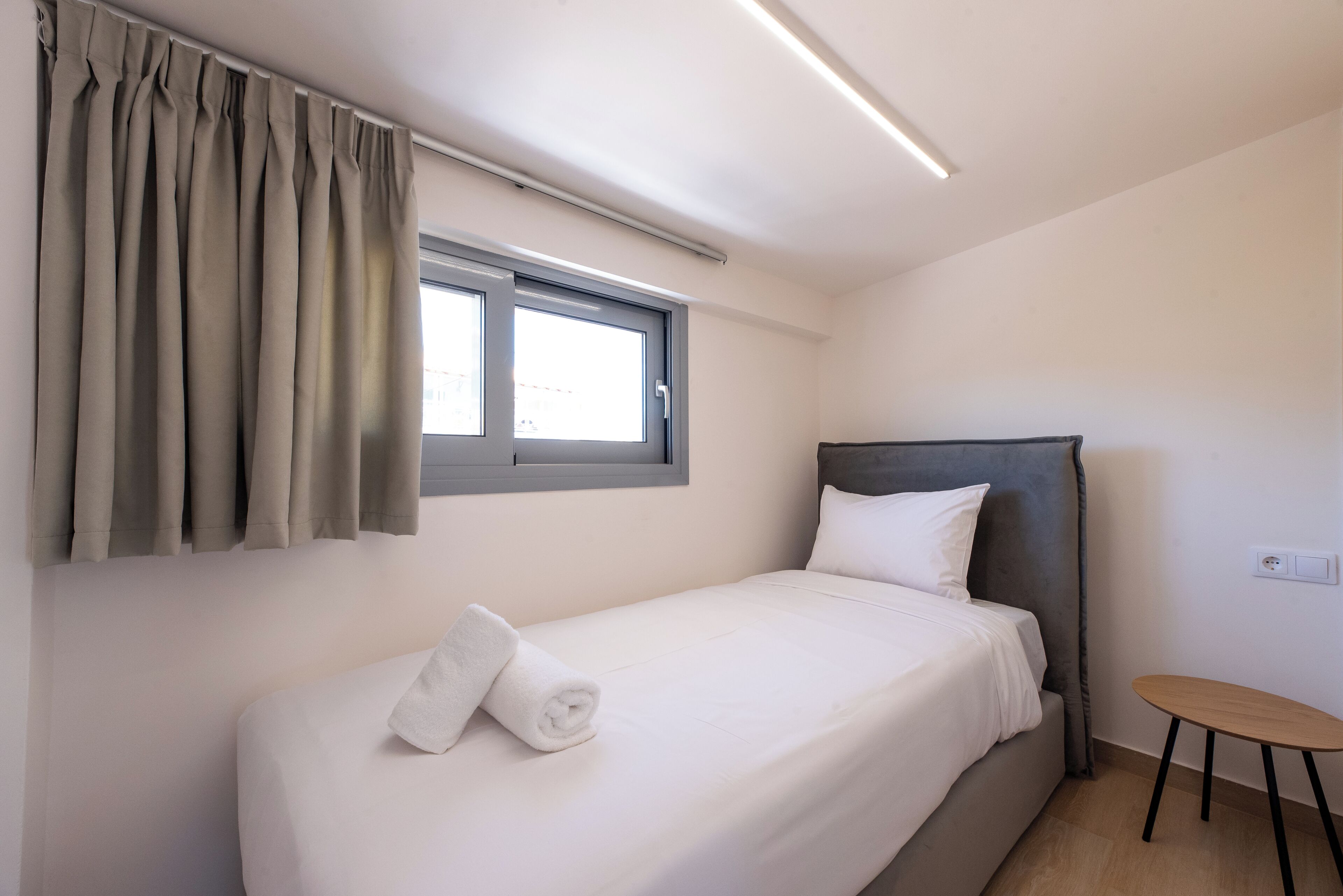 A single bed with a grey headboard, a small round table, a window with curtains, and a wall socket.