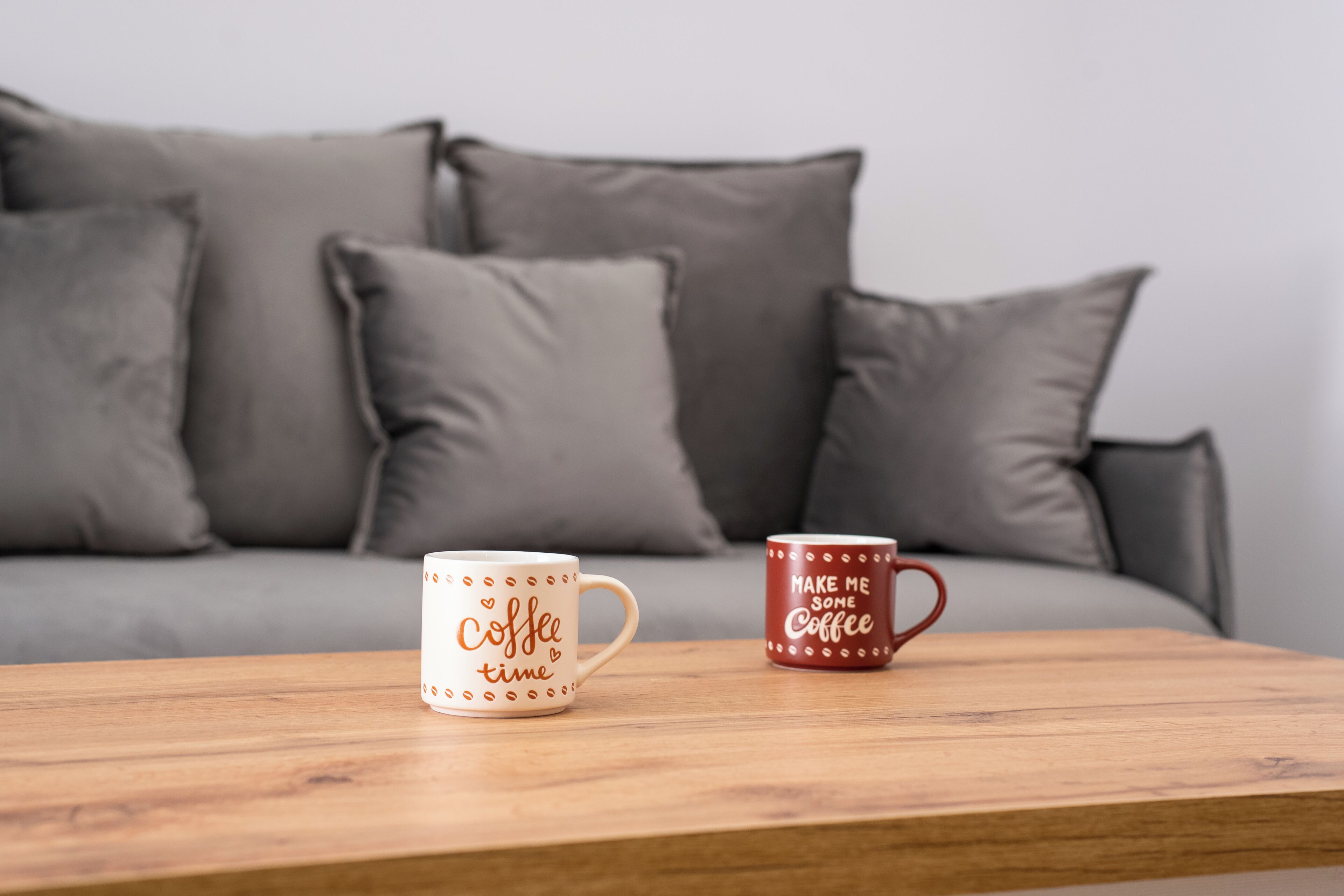 Twee koffiemokken op een houten tafel voor een grijze bank met kussens.