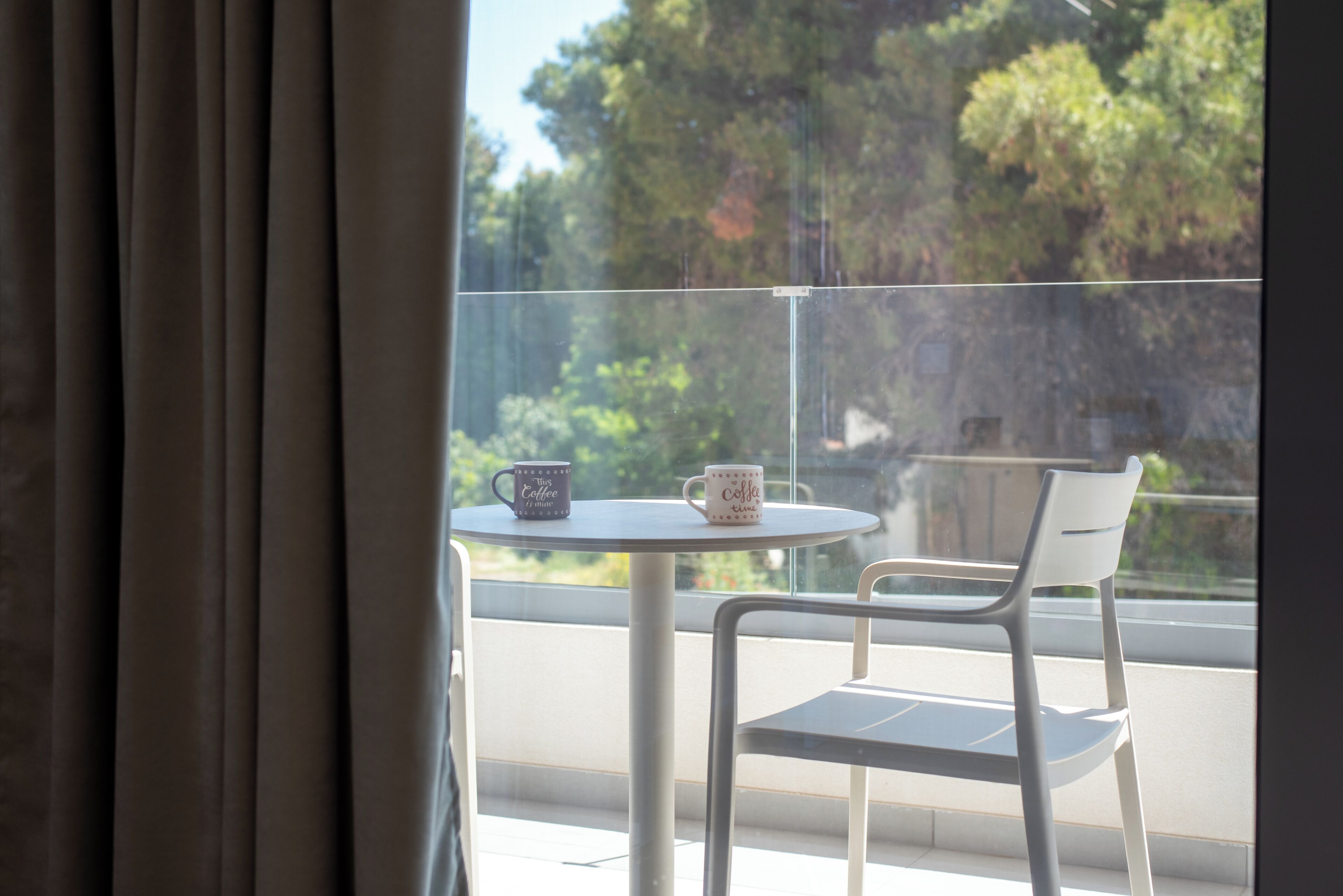 Un balcón con una mesa y dos sillas, una ventana con cortinas y vistas a árboles.