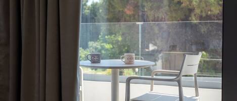 Un balcón con una mesa y dos sillas, una ventana con cortinas y vistas a árboles.