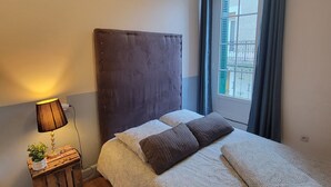 Une chambre à coucher avec un lit, une lampe de chevet, une petite table avec une plante et une fenêtre avec des rideaux.