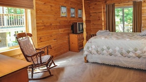 Una habitación de cabaña de madera con cama, sillón mecedor, televisor y ventilador de techo.