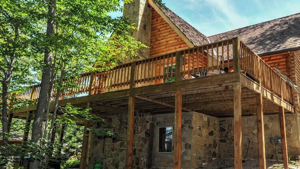 Una cabaña rústica con terraza de madera y paredes de piedra, rodeada de árboles.
