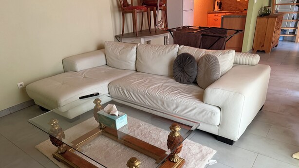 A living room with a white sofa, a glass coffee table with decorative items, and a kitchen in the background.