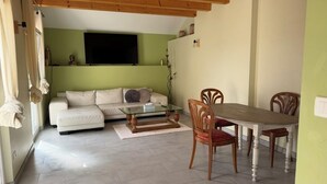 A living room with a sofa, a glass coffee table, wooden chairs, a dining table, and a TV mounted on the wall.