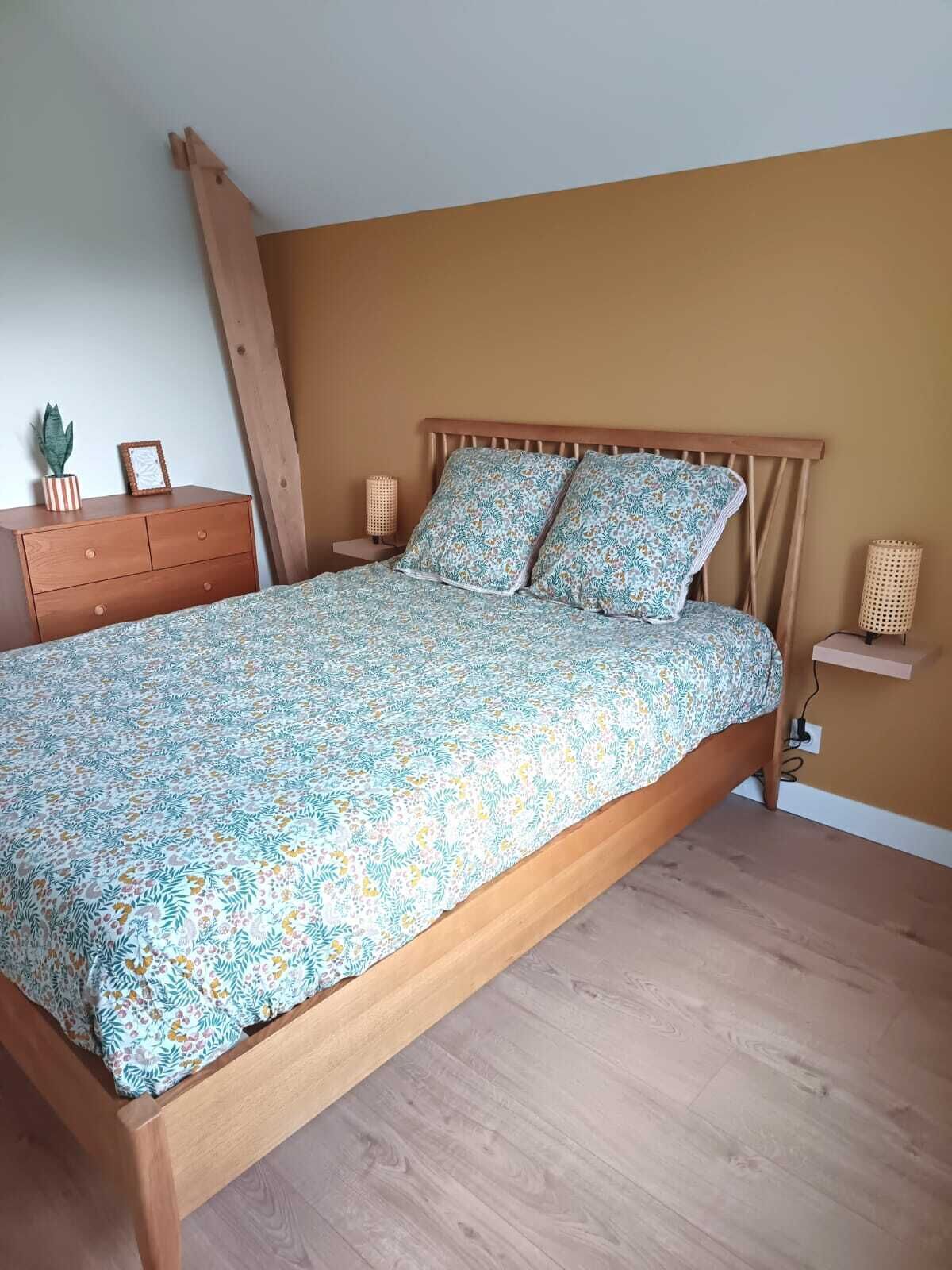 Quarto com cama de madeira, roupa de cama floral, mesa de cabeceira com luminária, cômoda e um vaso com planta.