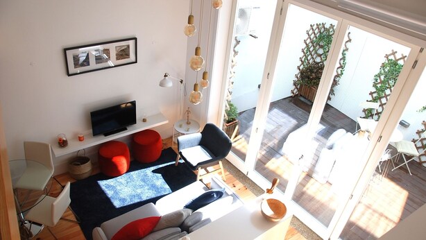 Un salon moderne doté d’un téléviseur à écran plat, de poufs rouges, d’une table à manger et d’une grande porte-fenêtre coulissante donnant sur une terrasse extérieure.