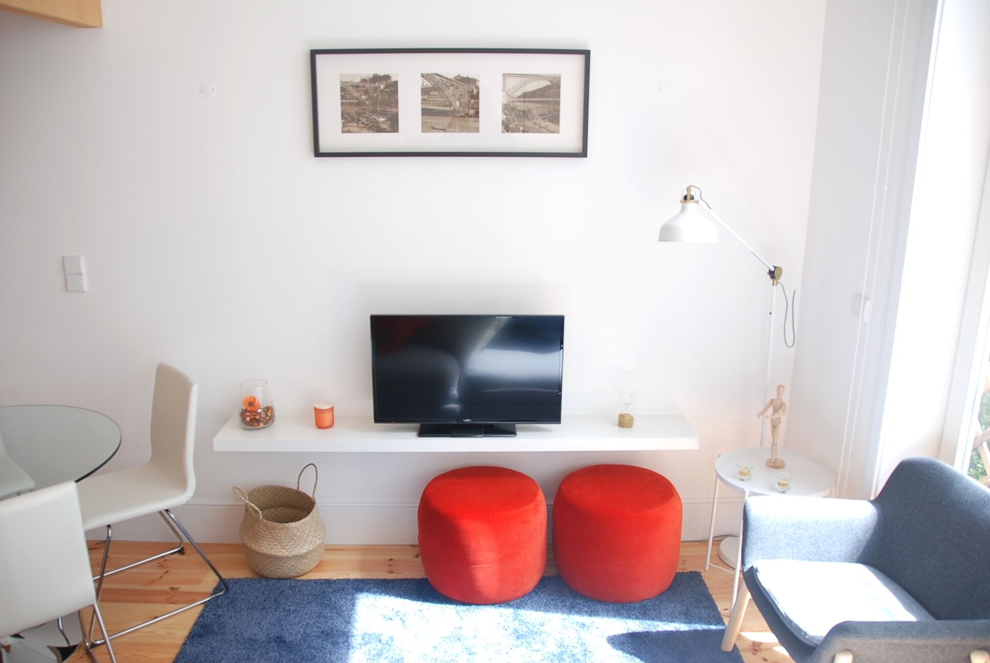 A modern living room with a flat-screen TV on a white shelf, two red ottomans, a glass coffee table, a white chair, and a small side table with a lamp.