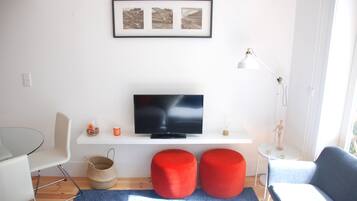 A modern living room with a flat-screen TV on a white shelf, two red ottomans, a glass coffee table, a white chair, and a small side table with a lamp.
