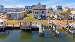 A coastal residential area with a prominent house featuring a porch and a dock extending into a calm body of water. - Canal Front Home, Boat Dock, Sleeps 17, Dog Friendly (Surf City)