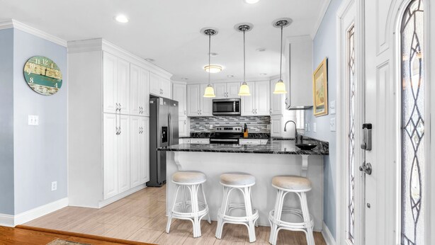 A modern kitchen with white cabinets, a stainless steel refrigerator, a microwave, and three white bar stools. - Canal Front Home, Boat Dock, Sleeps 17, Dog Friendly (Surf City)