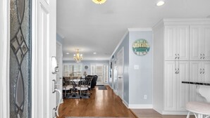 A spacious hallway with a dining area featuring a chandelier, a clock, and a kitchen area with white cabinets and a marble countertop. - Canal Front Home, Boat Dock, Sleeps 17, Dog Friendly (Surf City)
