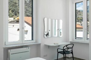 A room with a bed, a chair, a white wall-mounted mirror, and a window with a view of trees and buildings. - House in the center of Amalfi enveloped by the scent of lemons, far from the chaos (Amalfi)