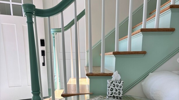 A glass-top table with a woven base, a book, and a tissue box on it, positioned near a staircase with teal railings and wooden steps.
