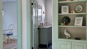A hallway with a light green door leading to a kitchen area. The kitchen has a white countertop, a sink, and a cabinet. There are decorative items on the shelves, including a bird figurine and a shell.
